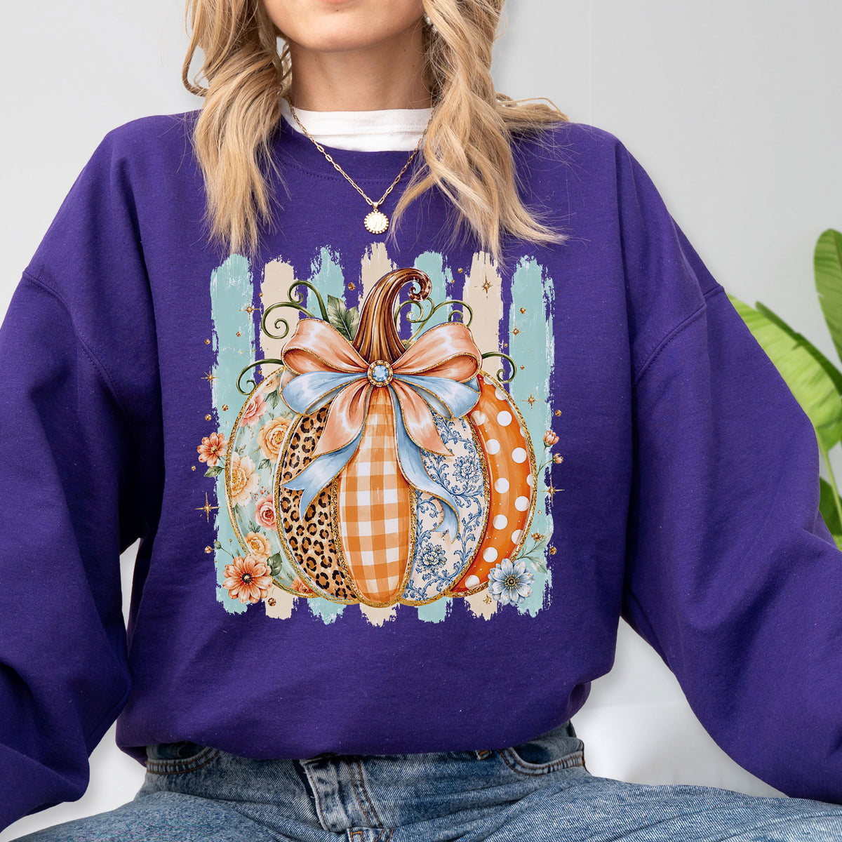 May include: A deep purple long-sleeve sweatshirt featuring a central graphic of a pumpkin decorated with various patterns including leopard print, orange gingham, blue floral, and white polka dots, topped with a large light blue bow.