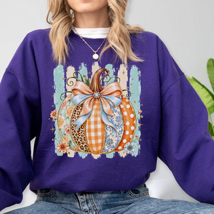 May include: A deep purple long-sleeve sweatshirt featuring a central graphic of a pumpkin decorated with various patterns including leopard print, orange gingham, blue floral, and white polka dots, topped with a large light blue bow.