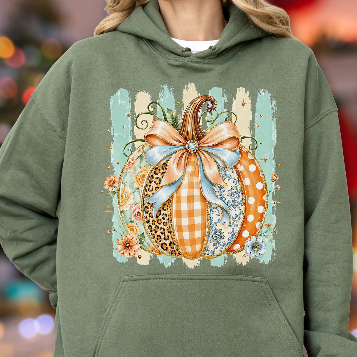 May include: An olive green hooded sweatshirt featuring a central graphic of a pumpkin decorated with various patterns including leopard print, orange gingham, floral motifs, and white polka dots, topped with a large light blue and peach bow.