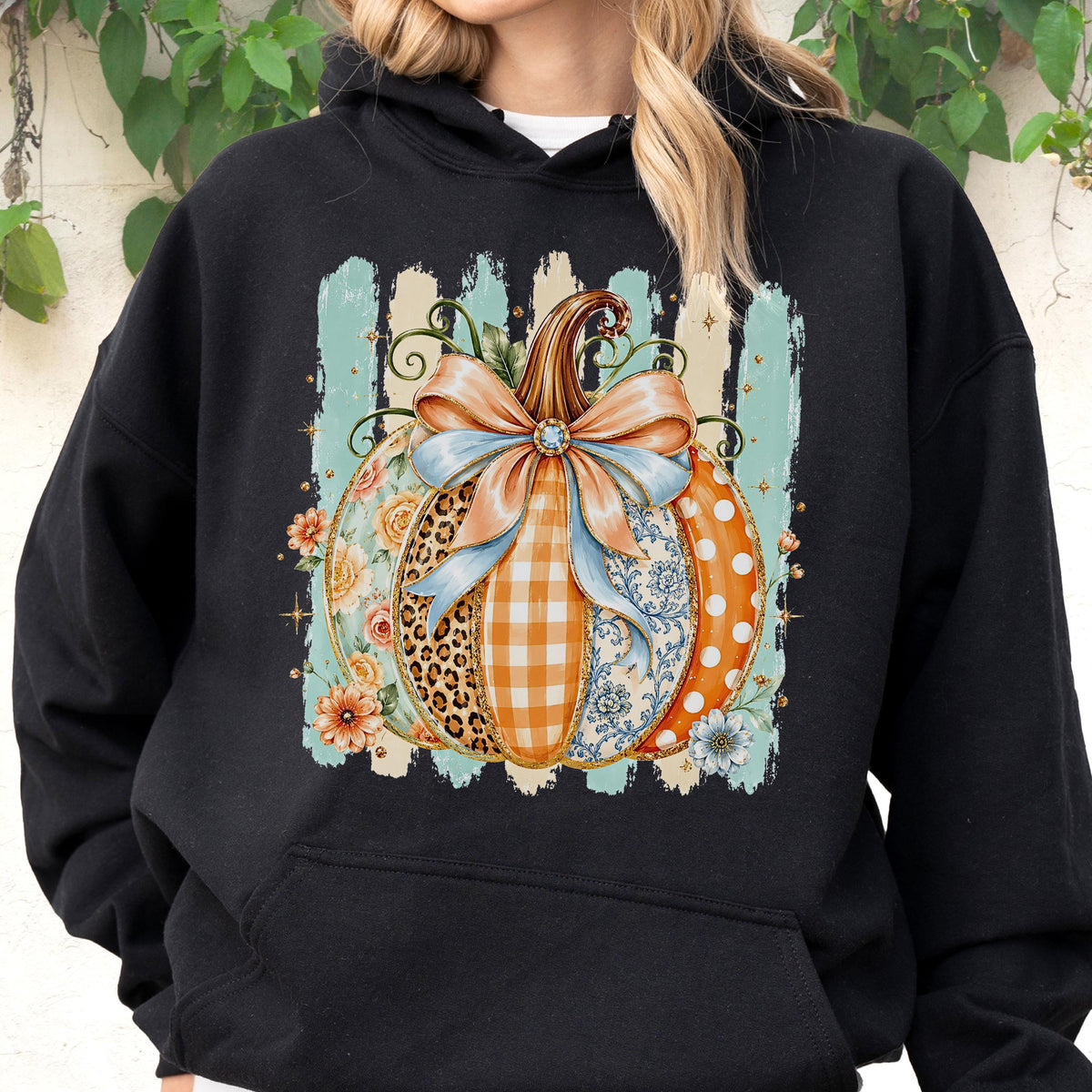 May include: A black hooded sweatshirt featuring a central graphic of a pumpkin decorated with various patterns including leopard print, orange gingham, and floral designs. The pumpkin is tied with a large light blue bow and surrounded by small flowers.