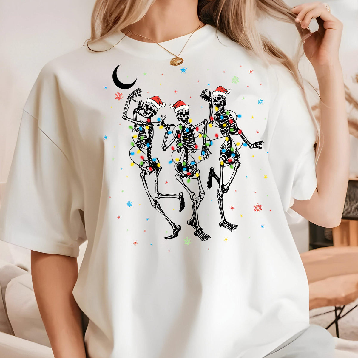 May include: A white short-sleeve t-shirt featuring a graphic of three skeletons wearing red Santa hats, wrapped in colorful Christmas light strings. The design includes a black crescent moon and scattered red, blue, and green star and snowflake icons.