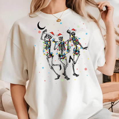 May include: A white short-sleeve t-shirt featuring a graphic of three skeletons wearing red Santa hats, wrapped in colorful Christmas light strings. The design includes a black crescent moon and scattered red, blue, and green star and snowflake icons.
