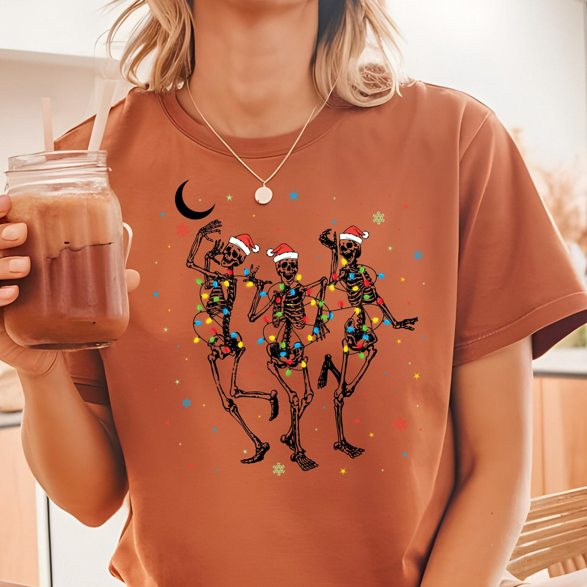 May include: A rust-colored t-shirt featuring a graphic of three black skeletons wearing red and white Santa hats. The skeletons are tangled in colorful Christmas lights, surrounded by small stars, snowflakes, and a black crescent moon design.