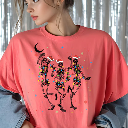 May include: A coral-colored crew neck t-shirt featuring a graphic of three dancing skeletons wearing red and white Santa hats. The skeletons are wrapped in colorful Christmas lights, accompanied by a black crescent moon and small snowflakes.