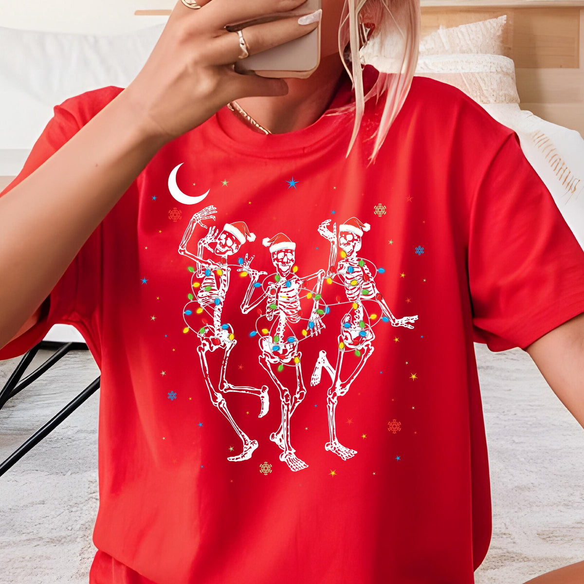 May include: A bright red short-sleeved t-shirt featuring a graphic of three white skeletons wearing Santa hats. The skeletons are tangled in colorful Christmas lights, surrounded by small white crescent moon, star, and snowflake icons.