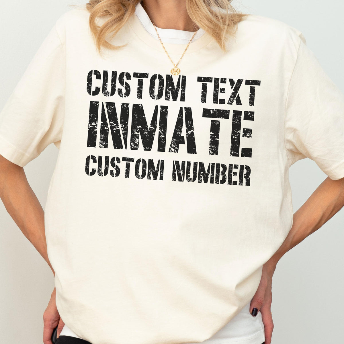 May include: A cream-colored short-sleeve t-shirt featuring black distressed-style text printed on the front. The text reads CUSTOM TEXT, INMATE, and CUSTOM NUMBER in a bold, blocky font. The shirt has a crew neckline and a relaxed fit.