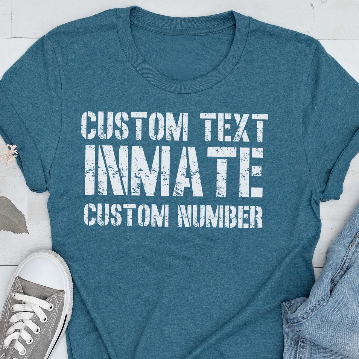 May include: A teal short-sleeved t-shirt featuring white distressed-style text that reads CUSTOM TEXT, INMATE, and CUSTOM NUMBER. The shirt has a crew neckline and is displayed alongside a gray sneaker and a portion of blue denim fabric.