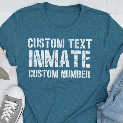 May include: A teal short-sleeved t-shirt featuring white distressed-style text that reads CUSTOM TEXT, INMATE, and CUSTOM NUMBER. The shirt has a crew neckline and is displayed alongside a gray sneaker and a portion of blue denim fabric.