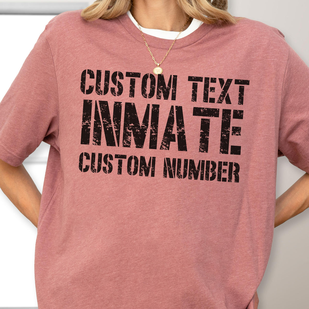 May include: A dusty rose short-sleeved t-shirt featuring black distressed-style lettering that reads CUSTOM TEXT, INMATE, and CUSTOM NUMBER. The shirt has a crew neckline and a heathered fabric texture, providing a casual and simple appearance.