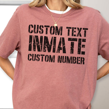 May include: A dusty rose short-sleeved t-shirt featuring black distressed-style lettering that reads CUSTOM TEXT, INMATE, and CUSTOM NUMBER. The shirt has a crew neckline and a heathered fabric texture, providing a casual and simple appearance.