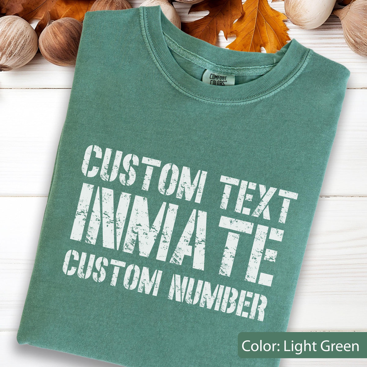 May include: A light green crew neck t-shirt featuring white distressed-style lettering that reads CUSTOM TEXT INMATE CUSTOM NUMBER. The garment has a Comfort Colors brand label visible at the inner neckline and a short-sleeved design.