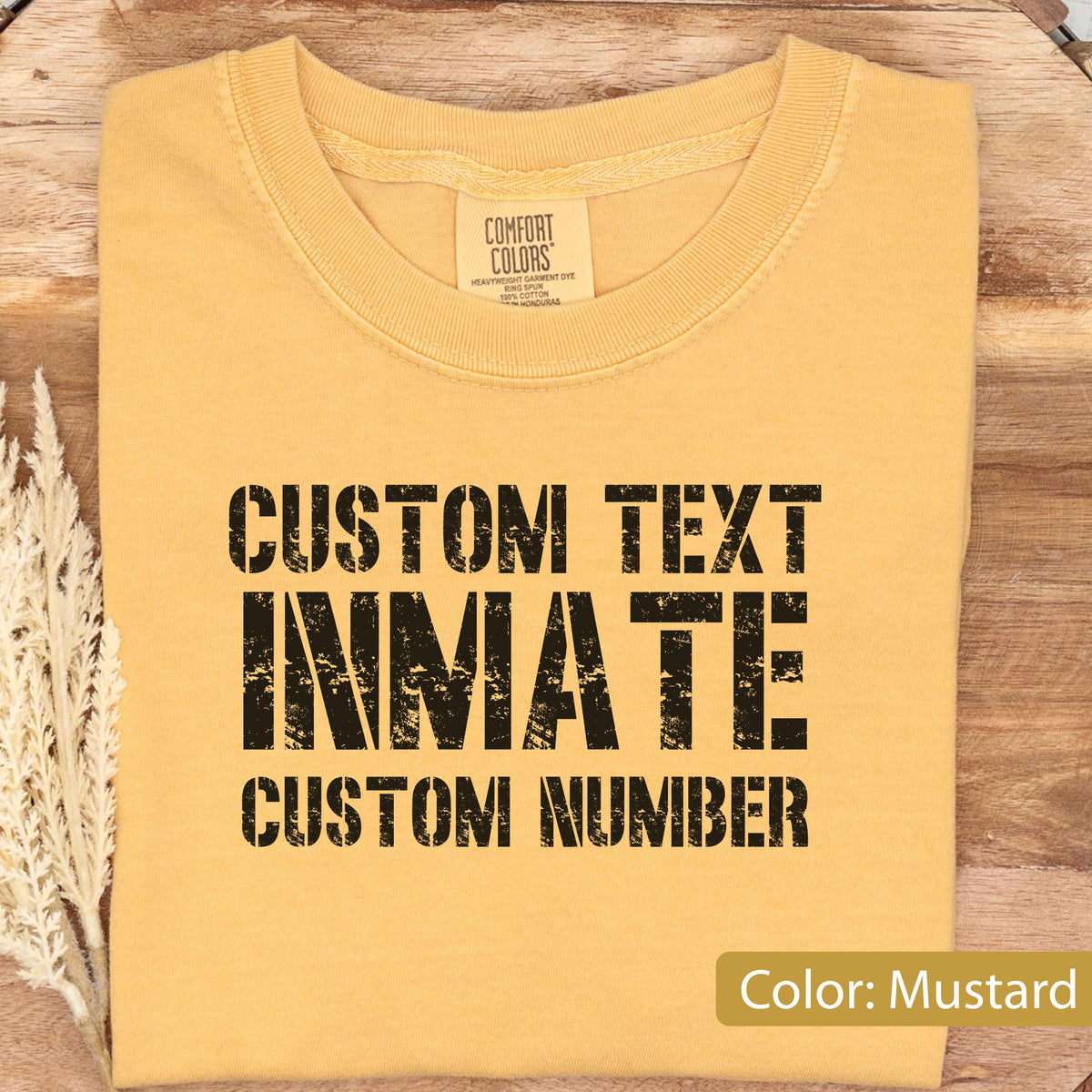 May include: A mustard yellow crew neck t-shirt with black distressed-style text printed on the front. The text reads CUSTOM TEXT, INMATE, and CUSTOM NUMBER. The shirt features a Comfort Colors brand label inside the collar area.