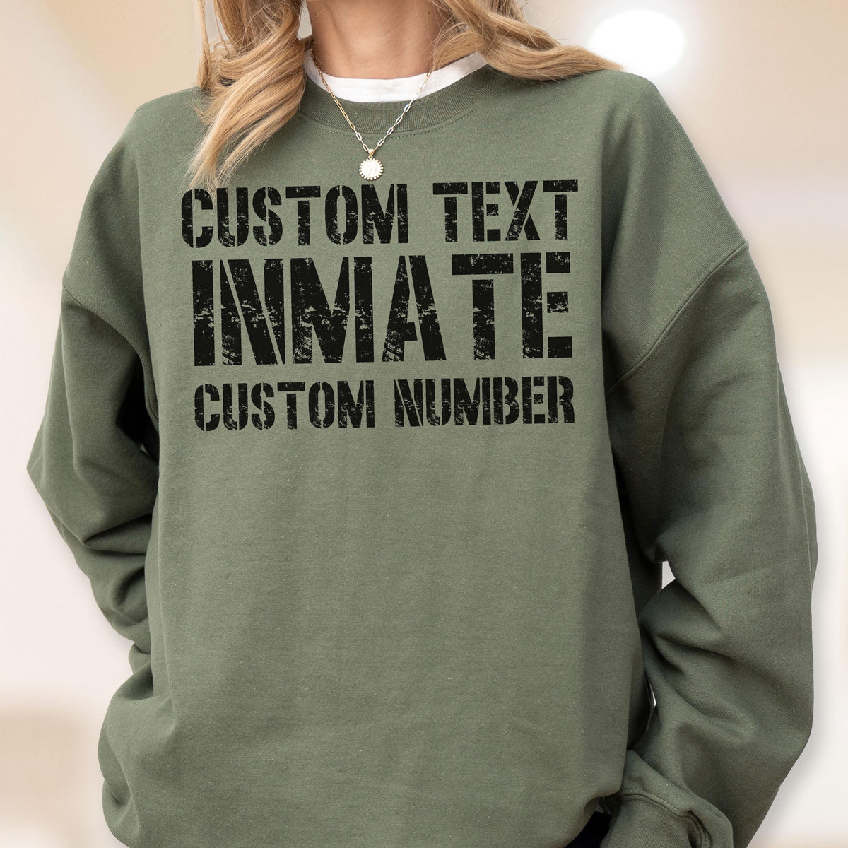 May include: An olive green crewneck sweatshirt featuring black distressed-style text printed on the chest. The text reads CUSTOM TEXT, INMATE, and CUSTOM NUMBER in a bold, block font. The garment has long sleeves and a ribbed collar neckline.