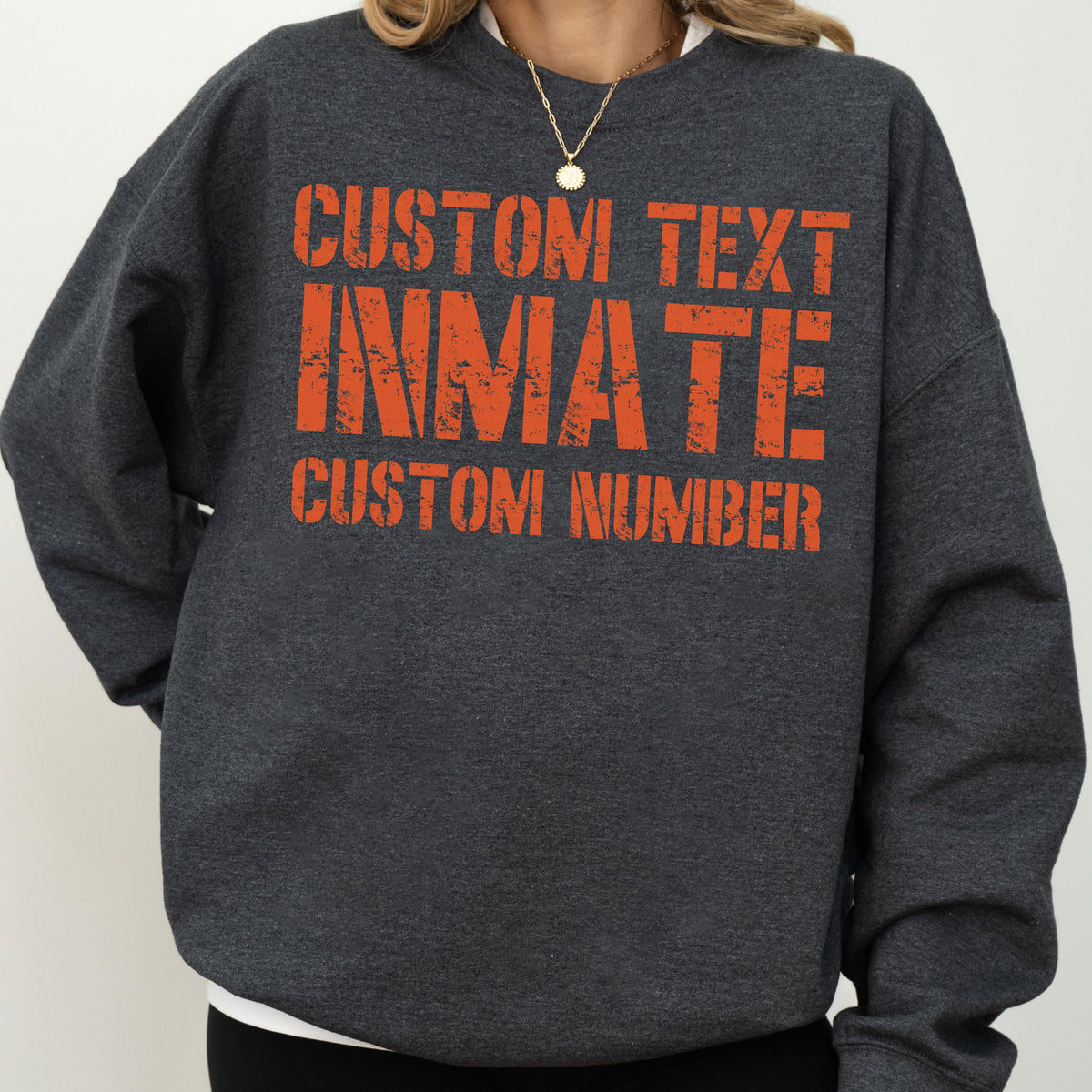 May include: A dark charcoal gray crewneck sweatshirt featuring orange distressed-style text on the chest. The graphic reads CUSTOM TEXT, INMATE, and CUSTOM NUMBER in a bold, blocky font. The garment has long sleeves and a ribbed neckline.