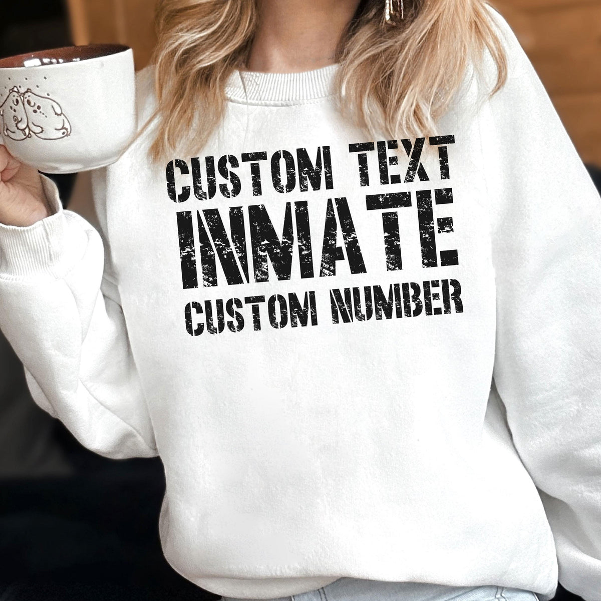 May include: A white crewneck sweatshirt featuring black distressed-style text that reads CUSTOM TEXT, INMATE, and CUSTOM NUMBER. The garment has long sleeves and a ribbed neckline, presented in a clean, simple design for casual wear.