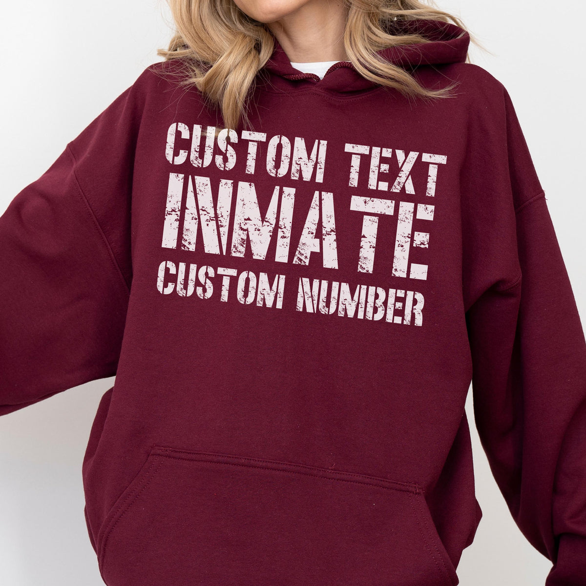 May include: A maroon hooded sweatshirt featuring white distressed-style text on the chest. The text reads CUSTOM TEXT, INMATE, and CUSTOM NUMBER in a stacked layout. The garment includes a front kangaroo pocket and long sleeves with ribbed cuffs.