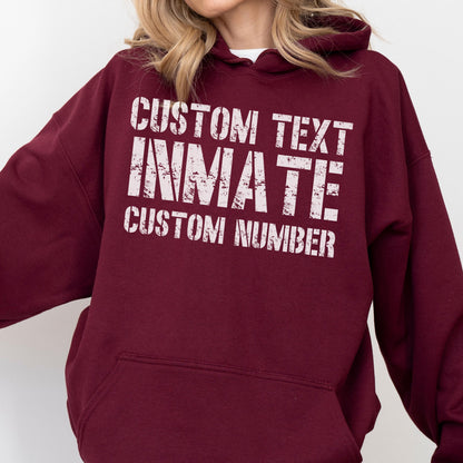 May include: A maroon hooded sweatshirt featuring white distressed-style text on the chest. The text reads CUSTOM TEXT, INMATE, and CUSTOM NUMBER in a stacked layout. The garment includes a front kangaroo pocket and long sleeves with ribbed cuffs.