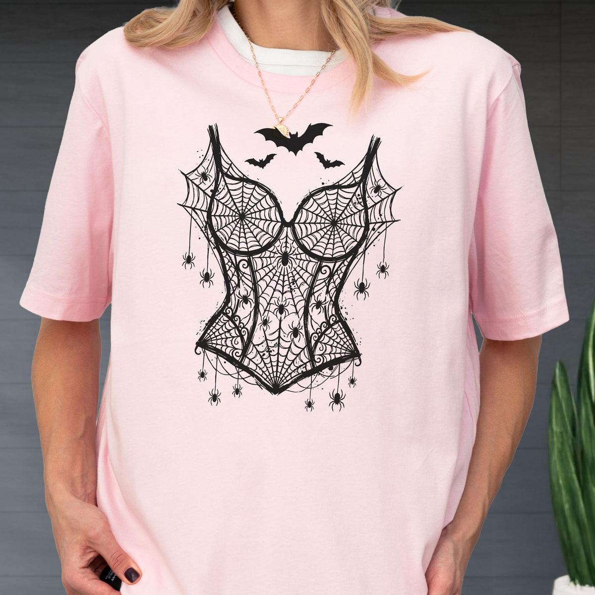 May include: A light pink short-sleeved t-shirt featuring a black graphic print of a corset made of spiderwebs. The design includes multiple small spiders hanging from the webbed corset and three black bats flying above the bodice area.
