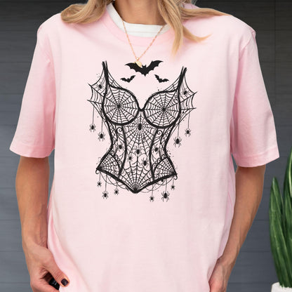 May include: A light pink short-sleeved t-shirt featuring a black graphic print of a corset made of spiderwebs. The design includes multiple small spiders hanging from the webbed corset and three black bats flying above the bodice area.