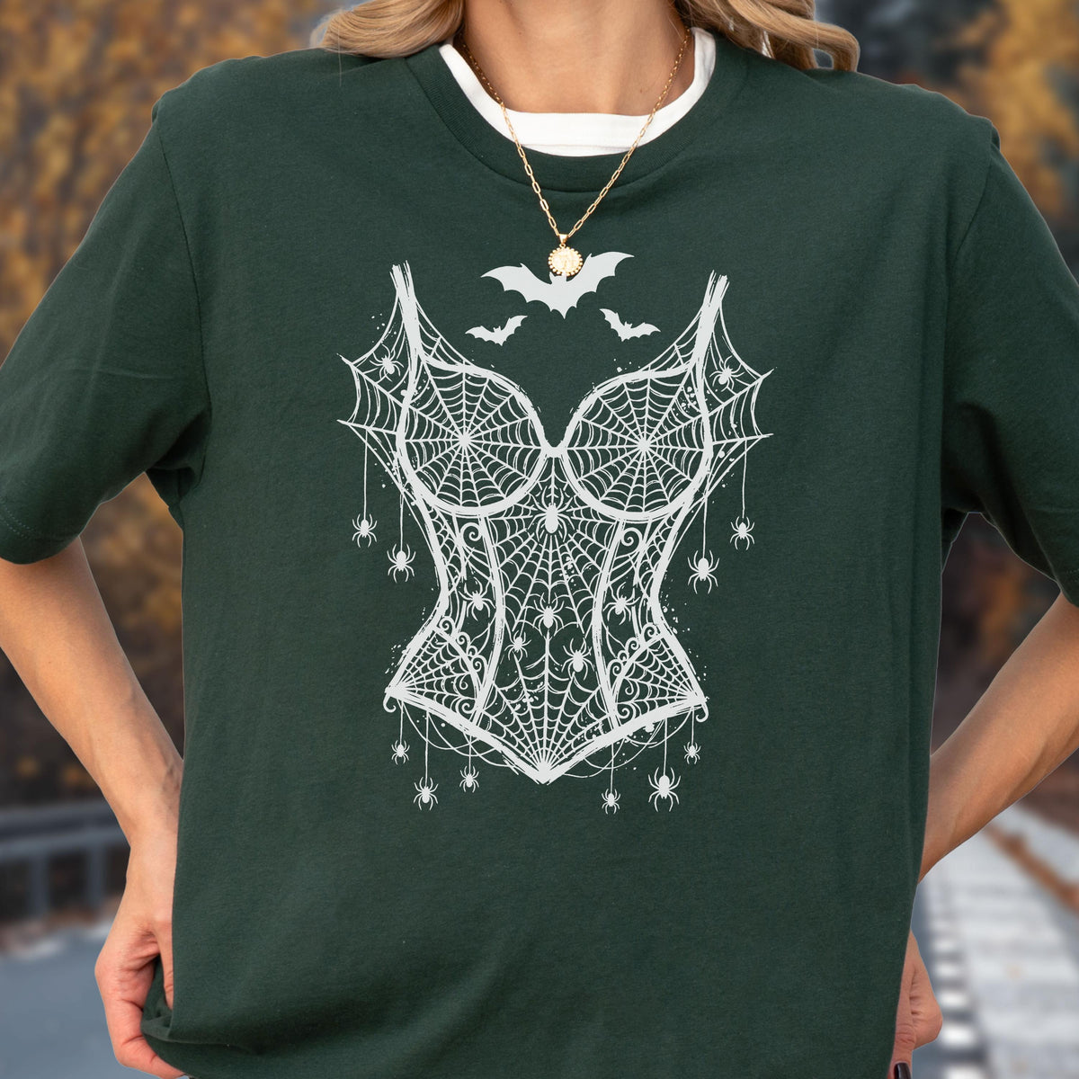 May include: A dark forest green short-sleeved t-shirt featuring a white graphic print of a corset made of spiderwebs, adorned with multiple small spiders and three bats flying above the neckline, worn with a simple gold-toned chain necklace.