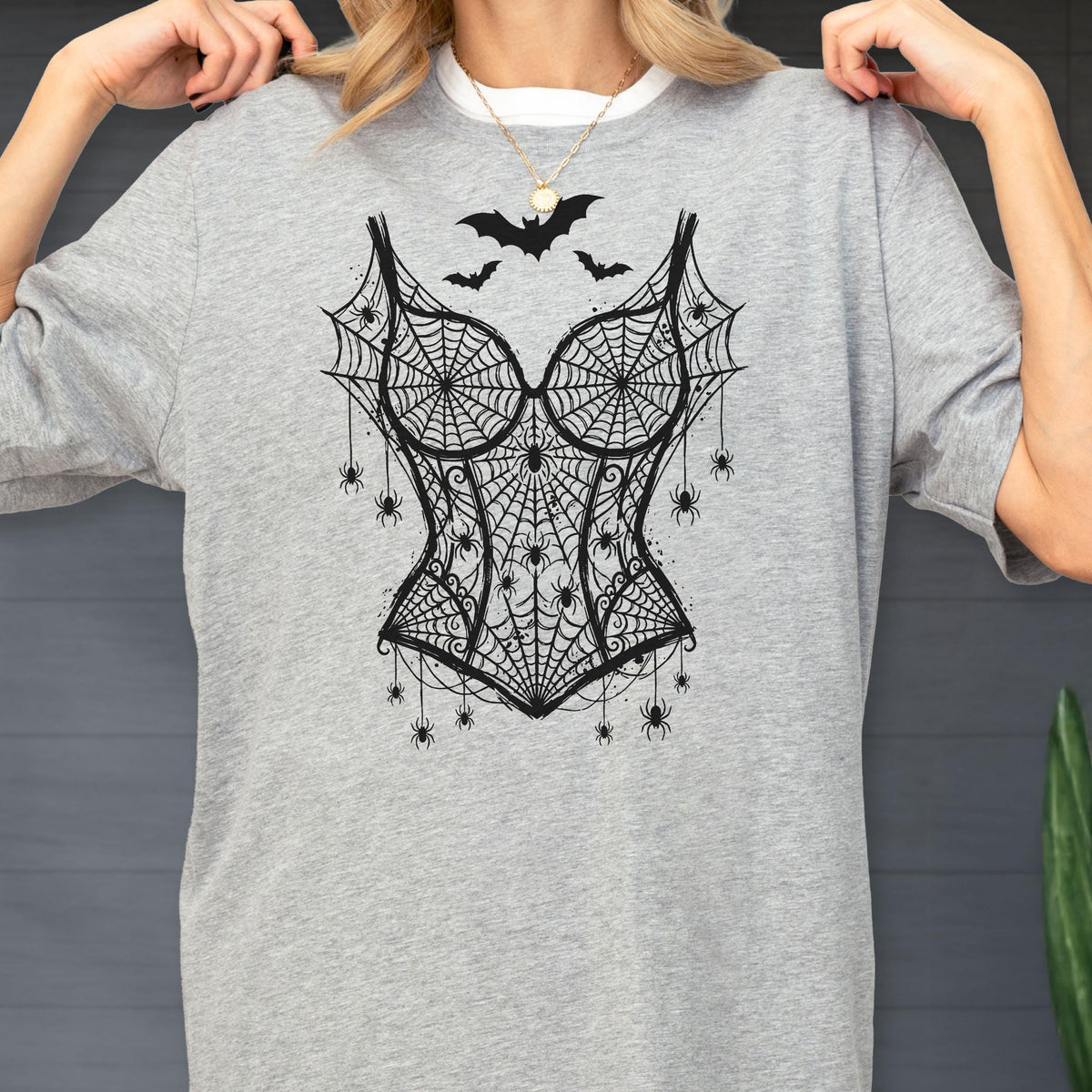 May include: A heather gray t-shirt featuring a black graphic print of a corset made from spiderwebs. The design includes multiple small spiders hanging from the webbed corset and three black bats flying above the neckline of the corset illustration.