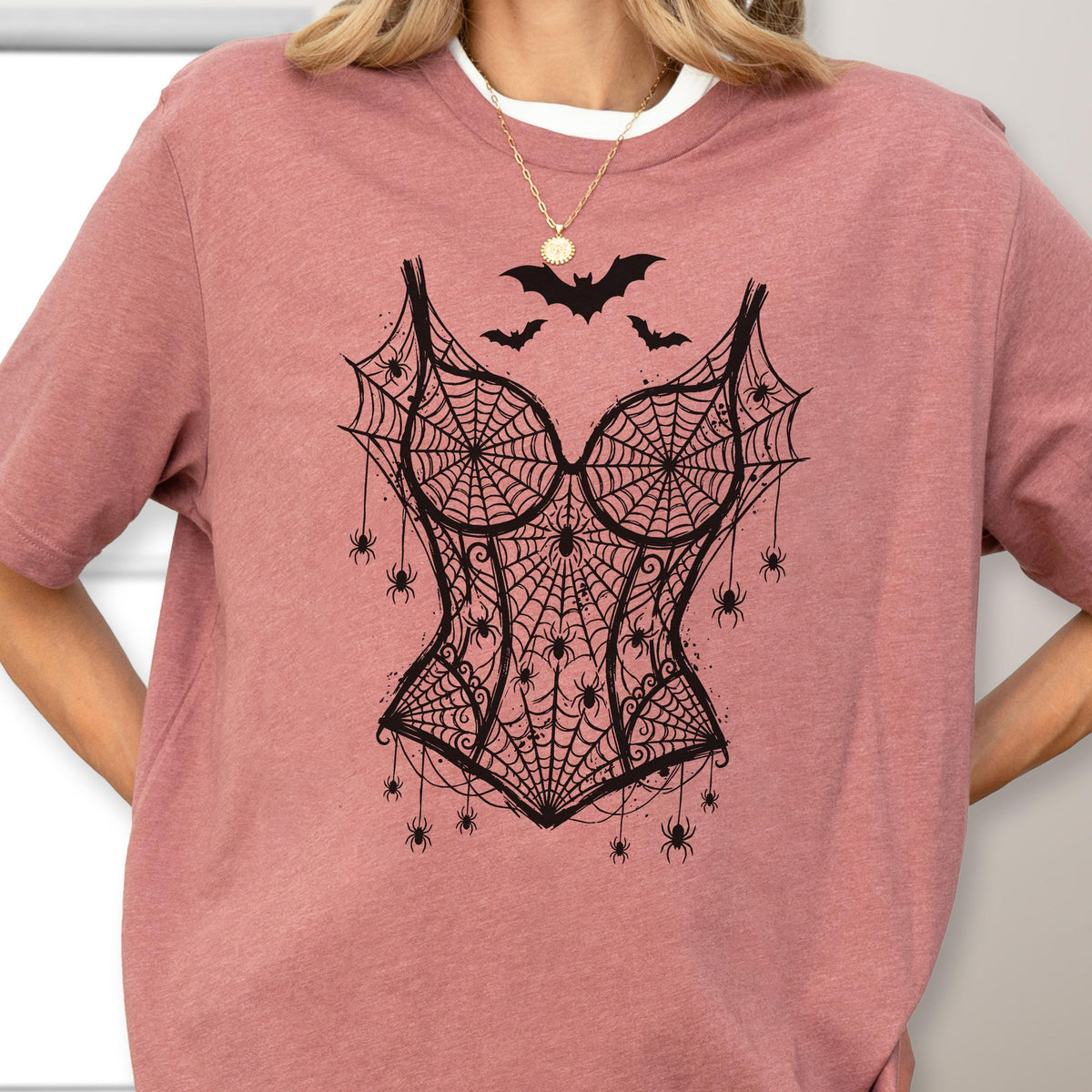 May include: A dusty rose short-sleeved t-shirt featuring a black graphic print of a corset made from spiderwebs. The design includes several small spiders hanging from the web pattern and three black bats flying above the corset graphic.
