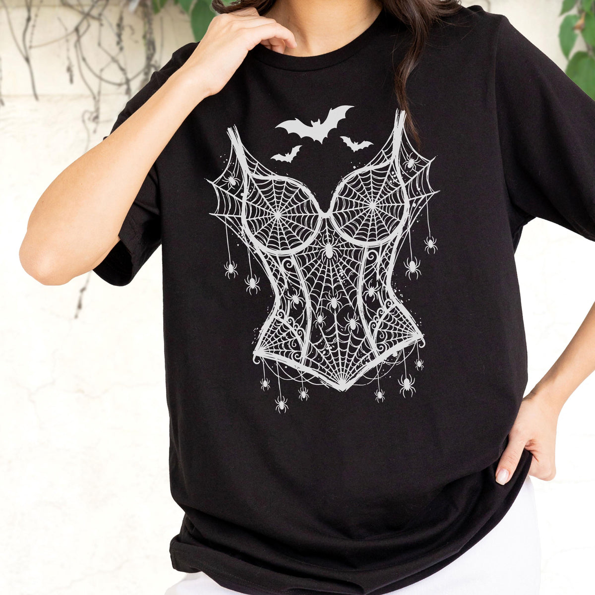 May include: A black short-sleeved t-shirt featuring a white graphic print of a corset design. The corset is composed of intricate spiderwebs and small spiders, with three bats flying above the neckline. The design covers the center chest area.
