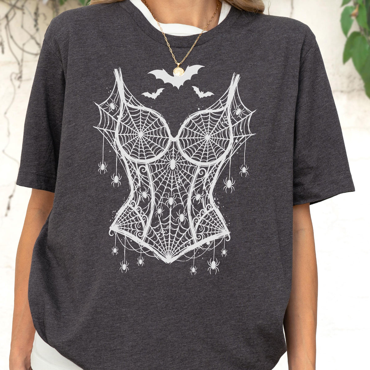 May include: A dark charcoal gray t-shirt featuring a white graphic print of a corset design. The corset is detailed with intricate spider web patterns, multiple small spiders hanging from the hem, and three bats flying above the neckline.