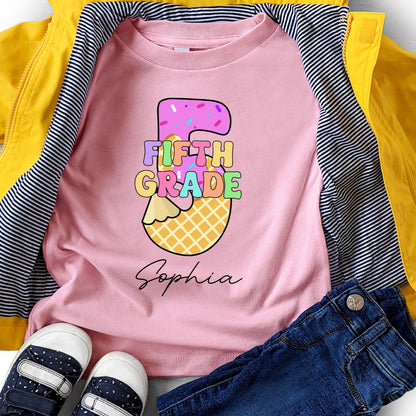 May include: Pink t-shirt with a colorful "Fifth Grade" graphic in a number 5 shape, decorated with sprinkles and a waffle cone base. The name "Sophia" is written in cursive below the graphic. The shirt is paired with blue jeans, a yellow jacket, and striped shirt.