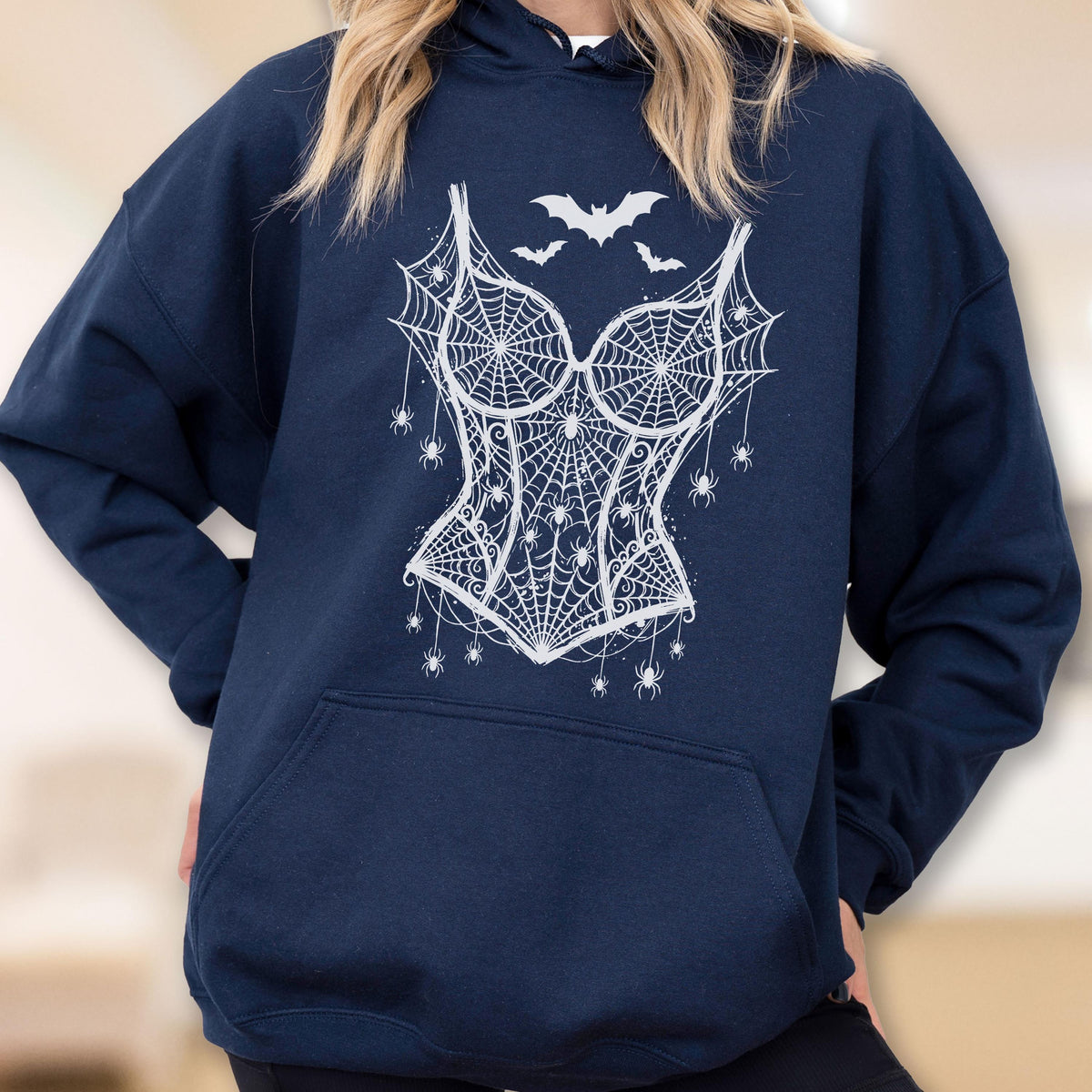 May include: A navy blue hooded sweatshirt featuring a white graphic print on the chest. The design depicts a corset shape formed by spiderwebs, accented with multiple small spiders hanging from threads and three bats flying above the corset.