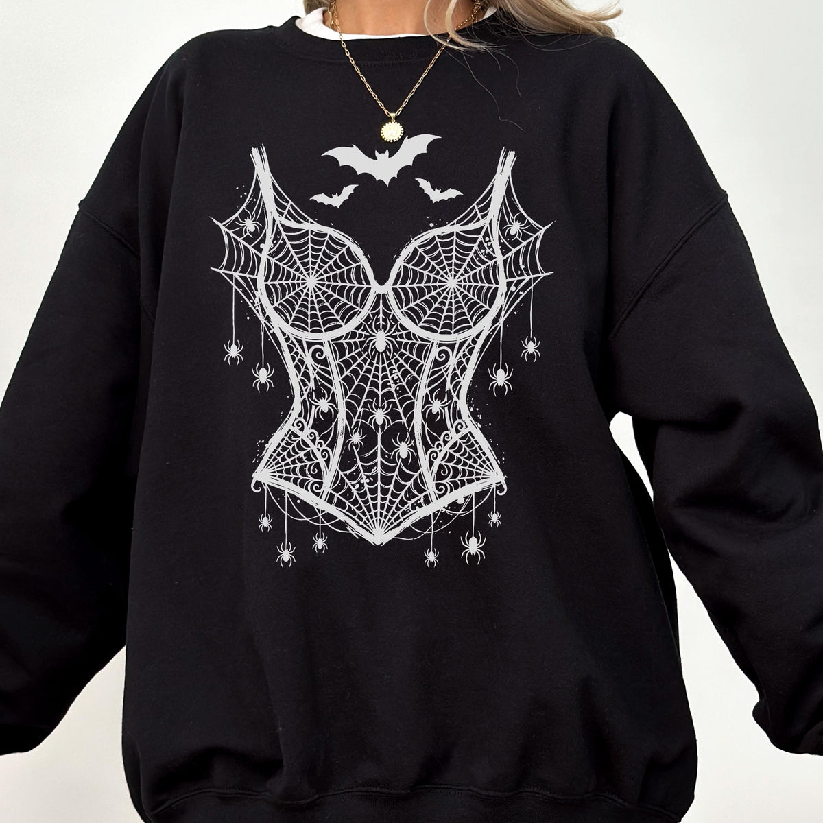 May include: A black long-sleeve sweatshirt featuring a white graphic print of a corset made from spiderwebs. The design includes several small spiders hanging from the webbed corset and three bats flying above the neckline of the corset graphic.