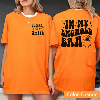 May include: An orange t-shirt featuring black text and graphics. The front displays "FUTURE MRS SMITH" with a small ring icon. The back features the large text "IN MY ENGAGED ERA" accompanied by star accents and a prominent diamond ring illustration.