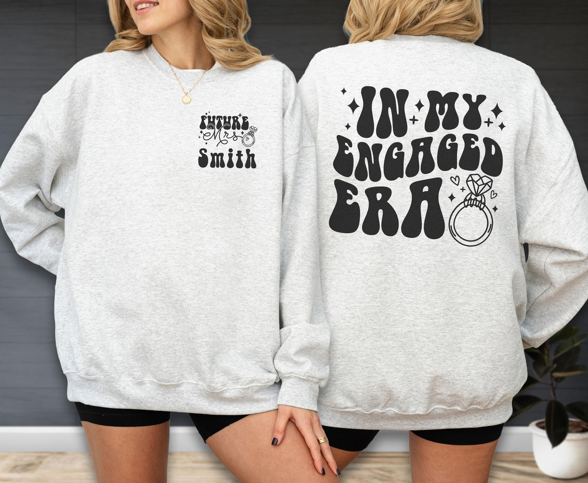 May include: A light gray crewneck sweatshirt featuring black text and graphics. The front left chest displays "Future Mrs. Smith" with a ring icon. The back features the text "IN MY ENGAGED ERA" in a bold, wavy font alongside a diamond ring graphic.