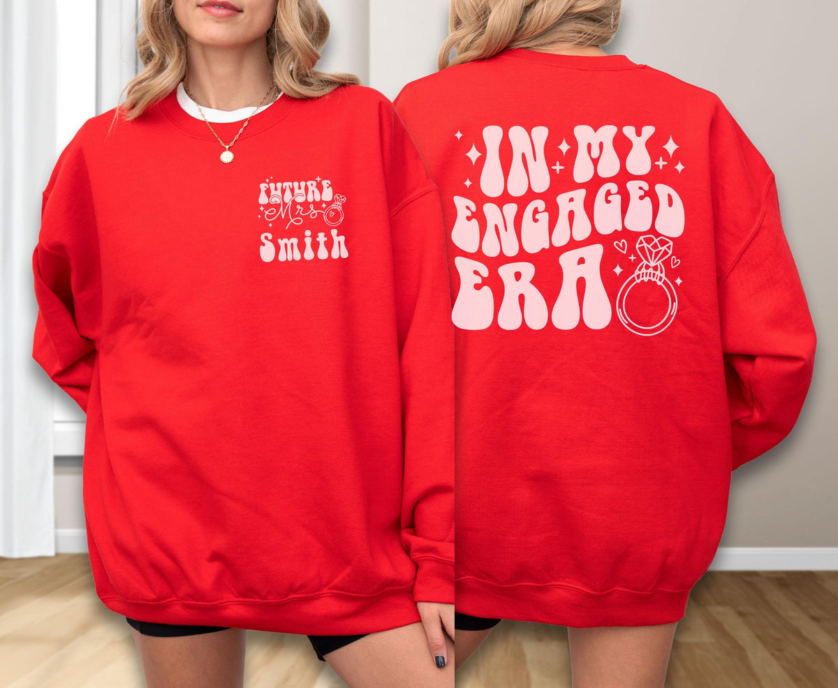 May include: A bright red crewneck sweatshirt featuring light pink graphic text. The front left chest displays "FUTURE MRS SMITH" with a small ring icon. The back features the large text "IN MY ENGAGED ERA" alongside a diamond ring illustration.