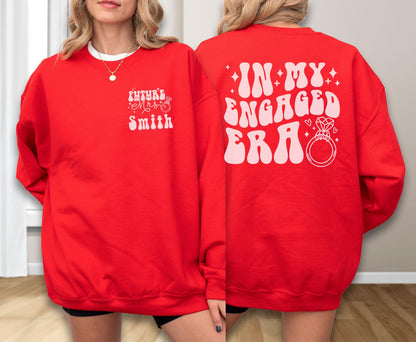 May include: A bright red crewneck sweatshirt featuring light pink graphic text. The front left chest displays "FUTURE MRS SMITH" with a small ring icon. The back features the large text "IN MY ENGAGED ERA" alongside a diamond ring illustration.