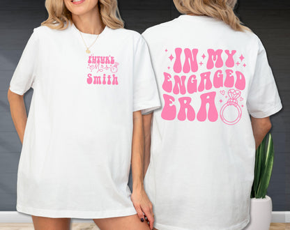 May include: A white short-sleeved t-shirt featuring pink text and graphics. The front displays "Future Mrs. Smith" with a ring icon. The back features the text "IN MY ENGAGED ERA" in large, bold pink letters, accompanied by a diamond ring graphic and stars.