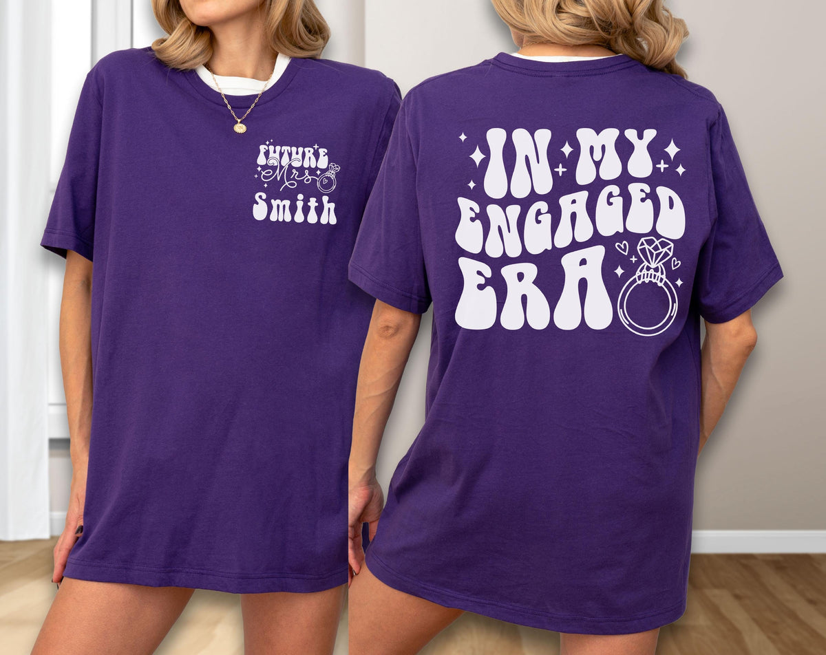 May include: A purple t-shirt featuring white text and graphics. The front left chest displays "FUTURE Mrs SMITH" with a ring icon. The back features the large white text "IN MY ENGAGED ERA" accompanied by a diamond ring illustration and small star accents.