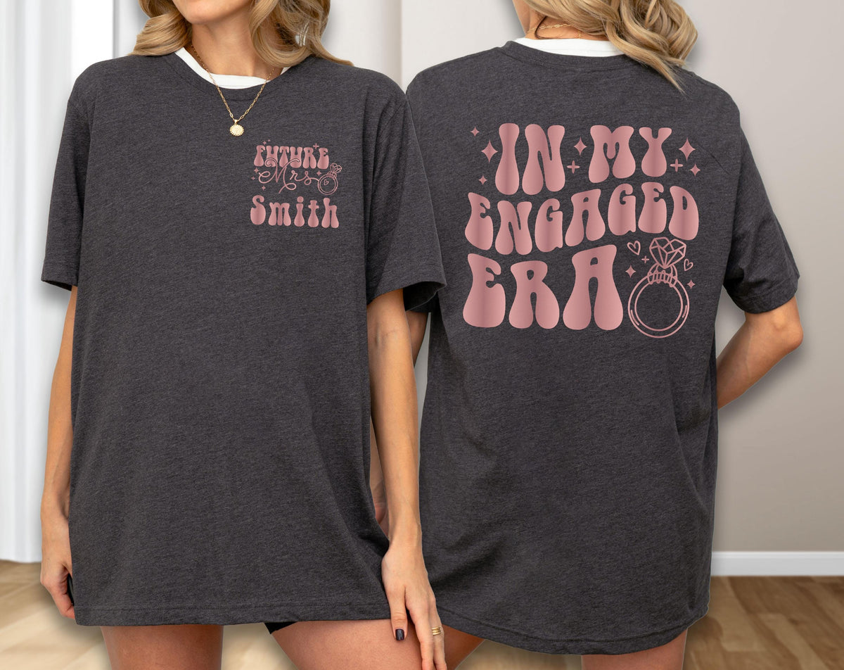 May include: A dark charcoal gray t-shirt featuring light pink text and graphics. The front left chest displays "Future Mrs. Smith" with a small ring icon. The back features the large text "IN MY ENGAGED ERA" with a diamond ring and star accents.