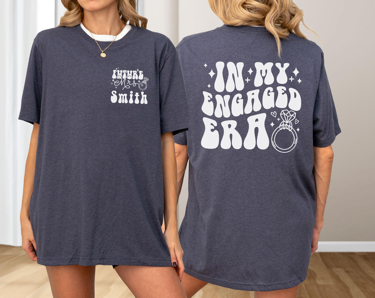 May include: A dark heather navy blue t-shirt featuring white screen-printed text. The front chest displays "Future Mrs. Smith" with a small ring icon. The back features the large text "IN MY ENGAGED ERA" in a bold, wavy font with a diamond ring graphic.