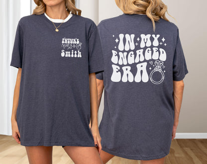 May include: A dark heather navy blue t-shirt featuring white screen-printed text. The front chest displays "Future Mrs. Smith" with a small ring icon. The back features the large text "IN MY ENGAGED ERA" in a bold, wavy font with a diamond ring graphic.