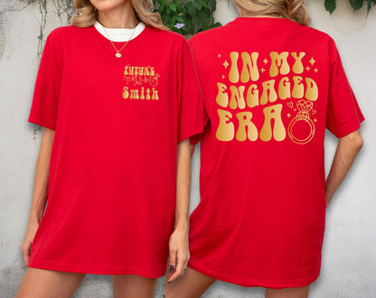 May include: A bright red short-sleeved t-shirt featuring gold text. The front displays "Future Mrs. Smith" with a small ring icon. The back features the large gold text "IN MY ENGAGED ERA" accompanied by a graphic of a diamond engagement ring.