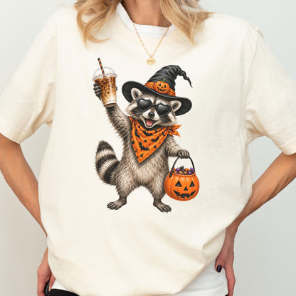 May include: A cream-colored t-shirt featuring a graphic of a raccoon wearing a black witch hat with a small pumpkin, heart-shaped sunglasses, and an orange bandana with black bats. The raccoon holds an iced coffee and a pumpkin candy bucket.