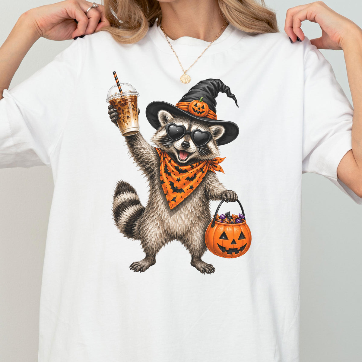 May include: A white t-shirt featuring a graphic of a raccoon wearing a black witch hat with a small pumpkin, heart-shaped sunglasses, and an orange bandana with a black bat pattern. The raccoon holds an iced coffee and a jack-o'-lantern candy bucket.