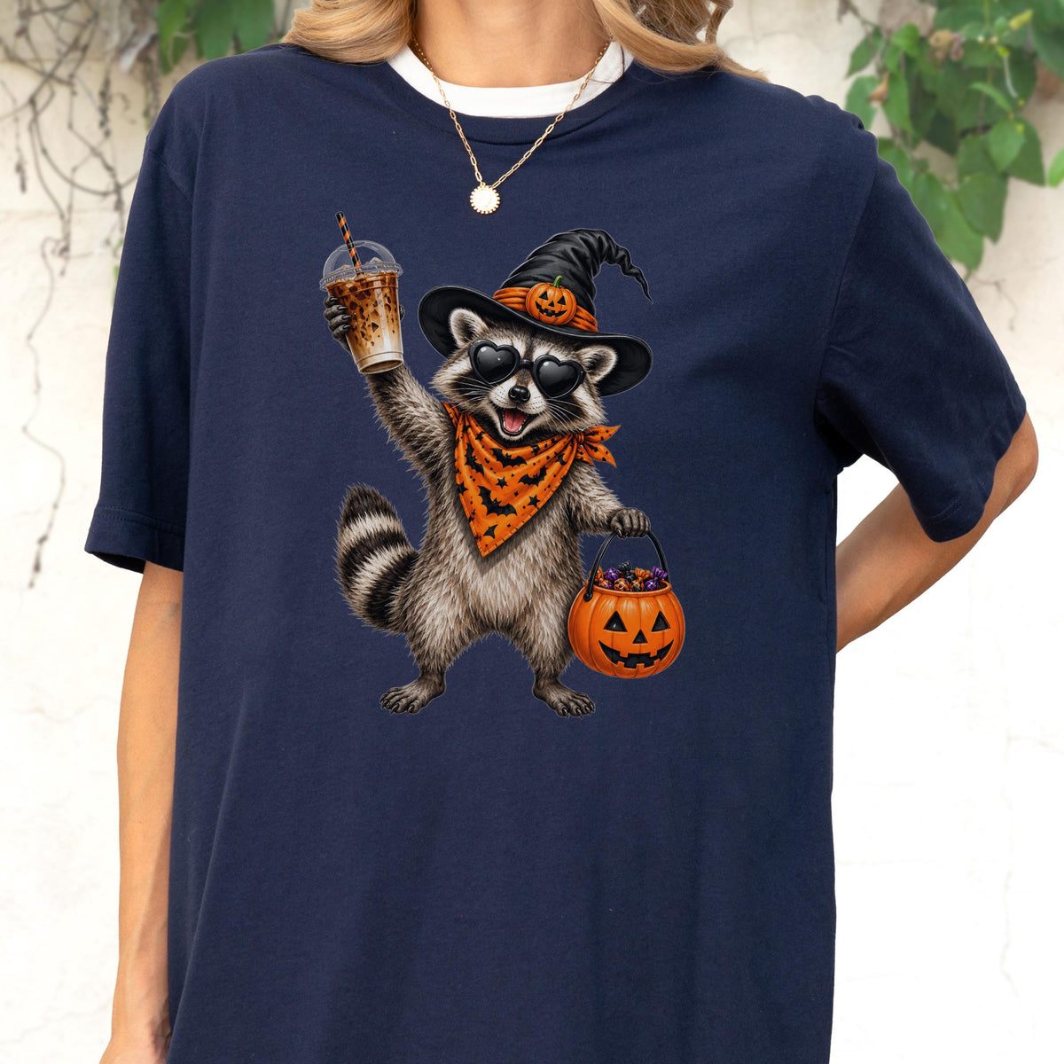 May include: A navy blue t-shirt featuring a graphic of a raccoon wearing a black witch hat with a small pumpkin, heart-shaped sunglasses, and an orange bandana with a black bat pattern. The raccoon holds an iced coffee and a pumpkin candy bucket.