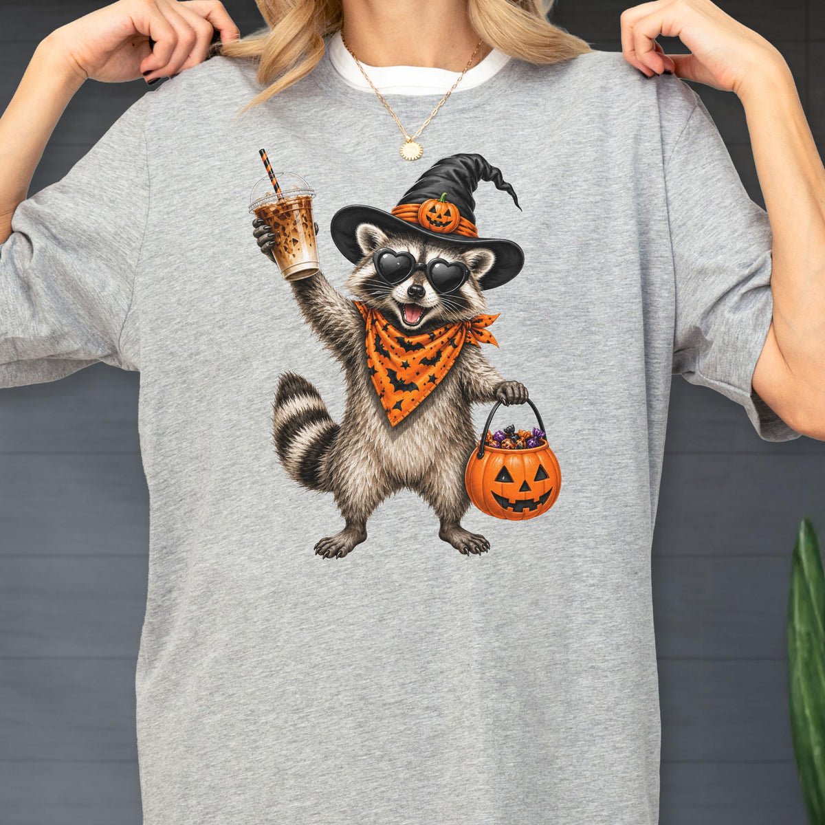 May include: A heather gray t-shirt featuring a graphic of a raccoon wearing a black witch hat with a pumpkin, heart-shaped sunglasses, and an orange bandana with a bat pattern. The raccoon holds an iced coffee and an orange jack-o'-lantern bucket.