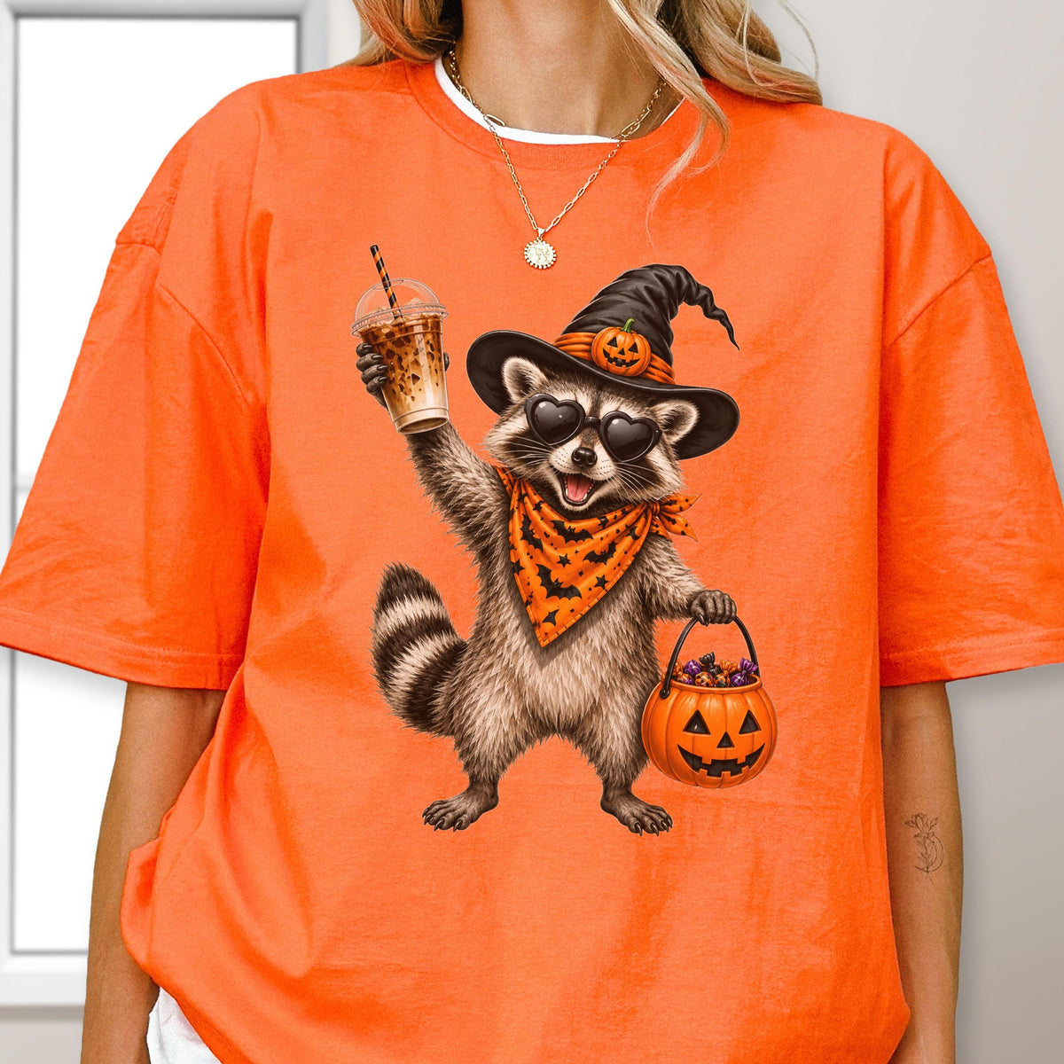 May include: An orange t-shirt featuring a graphic of a raccoon wearing a black witch hat with a small pumpkin, heart-shaped sunglasses, and an orange bandana with black bats. The raccoon holds an iced coffee and a jack-o'-lantern candy bucket.