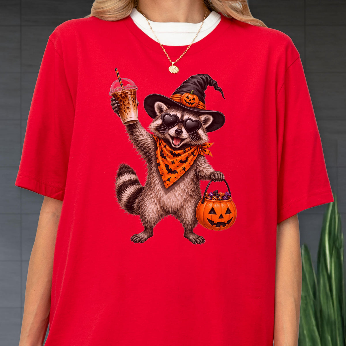 May include: A bright red t-shirt featuring a graphic of a raccoon wearing a black witch hat with a small pumpkin, heart-shaped sunglasses, and an orange bandana with a bat pattern. The raccoon holds a plastic cup and a jack-o'-lantern candy bucket.