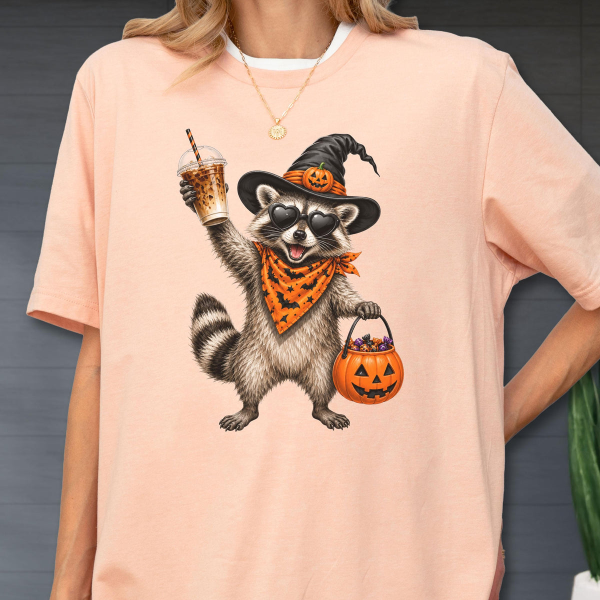 May include: A peach-colored t-shirt featuring a graphic of a raccoon wearing a black witch hat with a small pumpkin, heart-shaped sunglasses, and an orange bandana with a bat pattern. The raccoon holds an iced coffee and a pumpkin candy bucket.