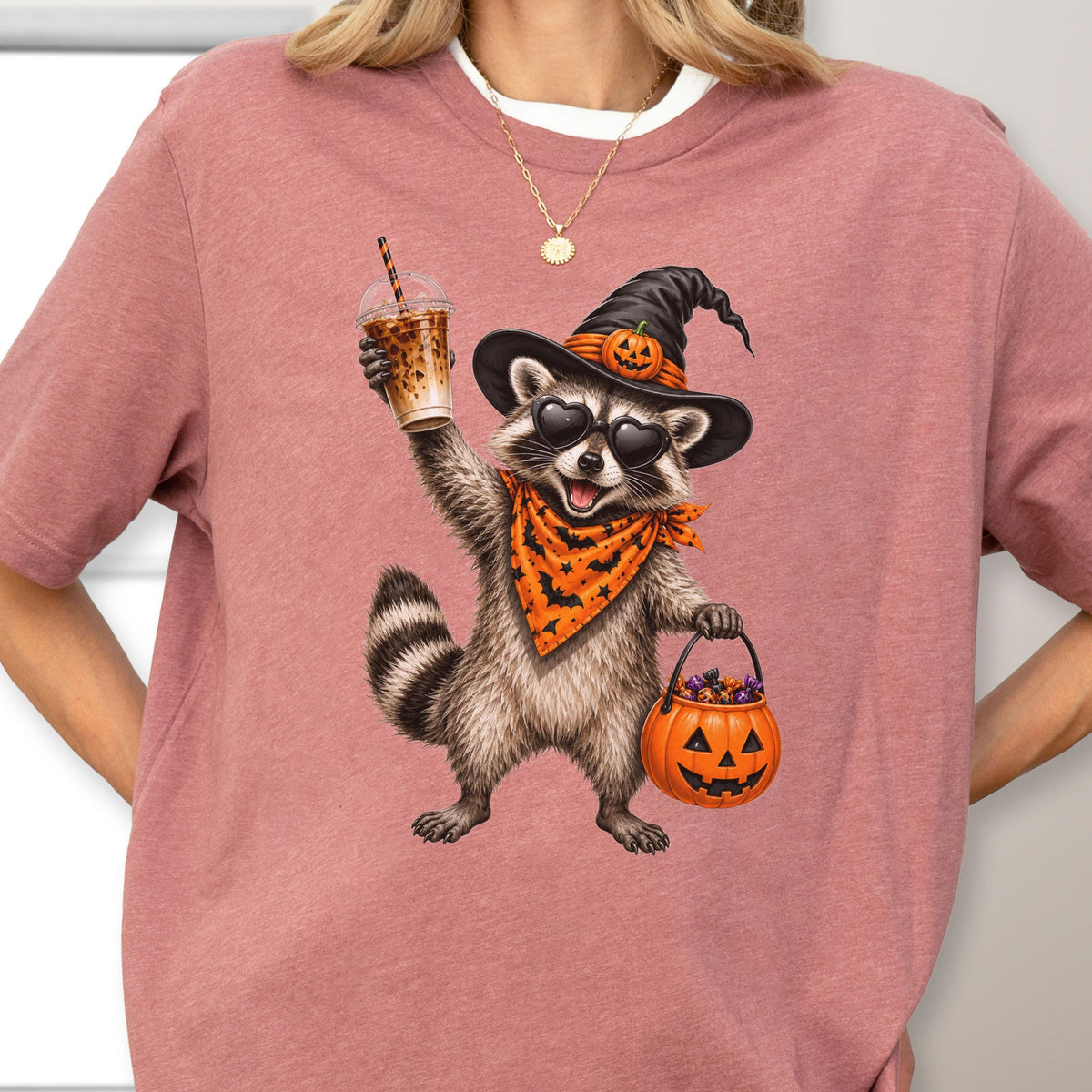 May include: A dusty rose t-shirt featuring a graphic of a raccoon wearing a black witch hat with a small pumpkin, heart-shaped sunglasses, and an orange bandana with black bats. The raccoon holds an iced coffee and a jack-o'-lantern candy bucket.
