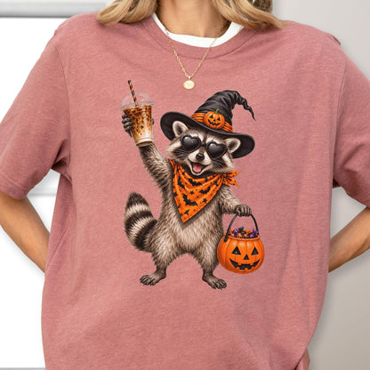 May include: A dusty rose t-shirt featuring a graphic of a raccoon wearing a black witch hat with a small pumpkin, heart-shaped sunglasses, and an orange bandana with black bats. The raccoon holds an iced coffee and a jack-o'-lantern candy bucket.